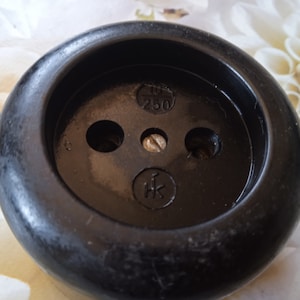 May include: Close-up of a vintage black electrical outlet. The outlet has a circular design with three holes and a central screw. The text "10 250" and a symbol are visible. The outlet is likely for electrical use.
