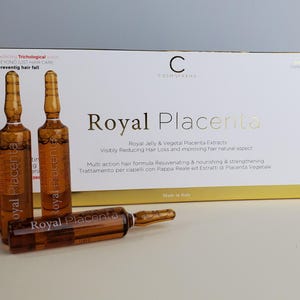以下が含まれることがあります： ロイヤルプラセンタのヘアトリートメント製品の箱。琥珀色のアンプルがいくつかあります。箱とアンプルには金色の文字が使われています。箱のテキストは、製品が抜け毛を減らし、髪の自然な状態を改善するためのものであることを示しています。