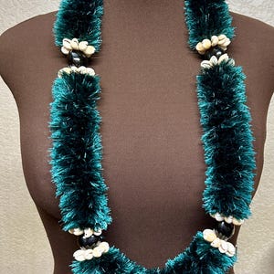 May include: A dark teal lei necklace with white accents and a white ribbon. The lei is made of a fluffy, textured material and features alternating sections of dark beads and white seashells. The necklace is displayed on a brown mannequin.