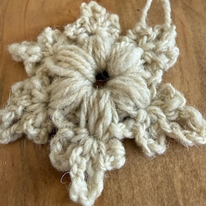 Può includere: Ornamento a forma di fiocco di neve all'uncinetto fatto a mano, color crema. Il fiocco di neve è realizzato con filato spesso, con un foro centrale e sei punte simmetriche. Un anello di filo è attaccato per appenderlo. L'ornamento è esposto su una superficie di legno.