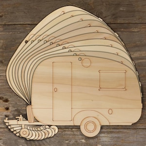 May include: A stack of light-colored wooden cutouts shaped like vintage travel trailers. Each piece features laser-etched outlines of a door, a rectangular window, a circular vent, and a wheel. The cutouts are arranged in a fanned-out pattern.