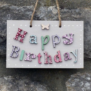 May include: A rectangular wooden sign with the words "Happy Birthday" in colorful, whimsical lettering. The letters are in various colors and patterns, with a small wooden butterfly and dotted border. The sign hangs by a jute rope.