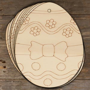 Könnte beinhalten: Ein Stapel unfertiger Holzzuschnitte in Eiform für Ostern. Jedes Ei hat geätzte Designs von Blumen, Wellenlinien, einer Schleife und Tupfen. Oben befindet sich ein kleines Loch zum Aufhängen. Die Eier sind aus hellem Holz.
