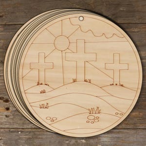 May include: A set of unfinished wooden round ornaments with a design of three crosses on a hill with a sun and clouds in the background. The ornaments are perfect for painting or staining.