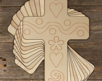 May include: A stack of unfinished wooden cross shapes with various designs, including hearts, swirls, and a flower. The crosses are ready to be painted or decorated.