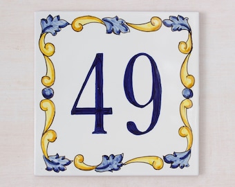May include: A white ceramic tile with a blue and yellow decorative border. The number 49 is painted in blue in the center of the tile.