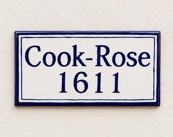 May include: A rectangular ceramic sign with a white background and a dark blue border. The text "Cook-Rose" is printed in dark blue on the top line, with the number "1611" printed in dark blue on the bottom line, centered within the frame.
