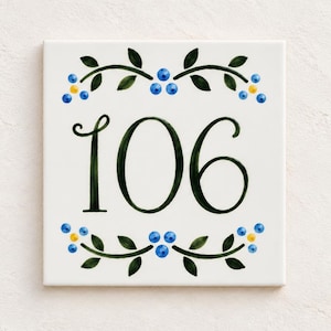 Könnte beinhalten: Eine quadratische weiße Keramikfliese mit der Hausnummer 106 in einer dunkelgrünen Schreibschrift. Das Design zeigt symmetrische dekorative Motive über und unter den Zahlen, bestehend aus dunkelgrünen Blättern, blauen Beeren und gelben Punkten.