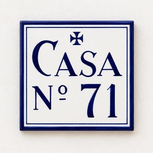Op de afbeelding: Een vierkante keramische tegel met een witte achtergrond en een donkerblauwe rand. De tekst "CASA No 71" is in het donkerblauw in het midden van de tegel gedrukt. Boven de tekst staat een klein donkerblauw kruissymbool. De tegel heeft een gladde afwerking.