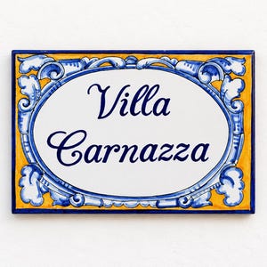 Könnte beinhalten: Ein rechteckiges Keramik-Hausschild mit einem weißen ovalen Zentrum und der dunkelblauen Aufschrift Villa Carnazza. Der Rand ist mit blauen Schnörkelmustern auf gelbem Hintergrund verziert und von einer dünnen dunkelblauen Kante umrahmt.