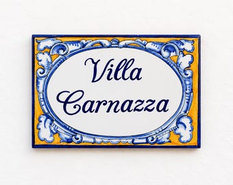 May include: A rectangular ceramic house sign featuring a white oval center with the dark blue text Villa Carnazza. The border is decorated with blue scrollwork patterns against a vibrant yellow background, framed by a thin dark blue edge.