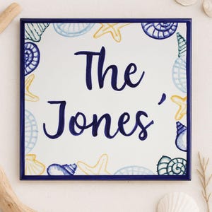 Può includere: Piastrella quadrata in ceramica con bordo blu navy e il nome "The Jones" in corsivo blu navy. La piastrella è decorata con conchiglie e stelle marine blu, gialle e bianche. Un oggetto decorativo per la casa.