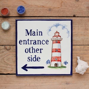 May include: A white ceramic tile with blue trim features a red and white striped lighthouse with a blue roof. The tile has the text "Main entrance other side" and an arrow pointing to the left.