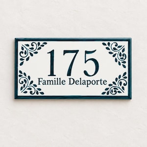 Könnte beinhalten: Ein rechteckiges Hausschild mit weißem Hintergrund und dunkelblauem Rand. Die Zahl 175 ist in großen dunkelblauen Ziffern in der Mitte aufgedruckt, darunter steht der Text Famille Delaporte. Dunkelblaue florale Ornamente verzieren die vier Ecken.