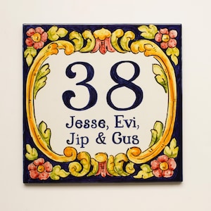 Könnte beinhalten: Quadratische Hausnummernkachel aus Keramik mit weißem Hintergrund, marineblauem Rand und dekorativem Blumen- und Schriftrollenmuster. Die Nummer "38" ist in Marineblau gehalten, darunter die Namen "Jesse, Evi, Jip & Gus".
