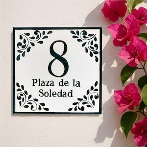 Op de afbeelding: Witte keramische huisnummerplaat met het nummer 8 en de tekst "Plaza de la Soledad" in het zwart. De plaat heeft een donkergroene rand en decoratieve hoekaccenten. Heldere roze bloemen op de achtergrond.