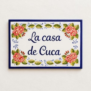 Könnte beinhalten: Ein rechteckiges Keramikschild mit dunkelblauem Rand und weißem Hintergrund. Der Text "La casa de Cuca" ist mittig in dunkelblauer Schrift zu sehen. Die Ecken sind mit handgemalten rosa Blumen und grünen Blättern verziert.