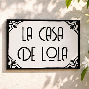 Könnte beinhalten: Ein rechteckiges Wandschild mit weißem Hintergrund und einem schwarzen Rand, der dekorative Eckmuster aufweist. Der Text LA CASA DE LOLA ist in einer fetten, schwarzen, stilisierten Schrift in der Mitte des Schildes mit schwarzem Rahmen gedruckt.