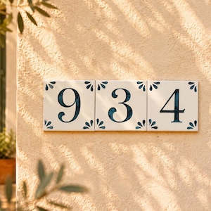 May include: Three square ceramic tiles arranged horizontally, each featuring a dark blue painted number on a white background. The tiles display the sequence 9, 3, and 4, with small blue floral-like corner accents on each individual tile piece.
