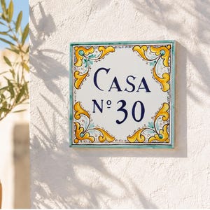 Op de afbeelding: Vierkant keramisch huisnummerbord met het woord "CASA" en het nummer "30" in marineblauw. Het bord heeft een witte achtergrond met een turquoise rand en gele en turquoise bloemenhoekontwerpen. Het bord is op een witte getextureerde muur gemonteerd.