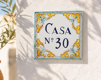 May include: Square ceramic house number sign with the word "CASA" and the number "30" in navy blue. The sign has a white background with a teal border and yellow and teal floral corner designs. The sign is mounted on a white textured wall.