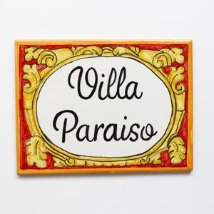 Könnte beinhalten: Eine rechteckige Wandplakette aus Keramik mit einem weißen ovalen Zentrum und dem schwarzen Text Villa Paraiso. Die Plakette ist von einem dekorativen gelb-goldenen Rand mit kunstvollen Schnörkeln auf rotem Hintergrund umgeben.