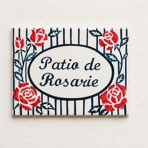 Op de afbeelding: Een rechthoekig keramisch bord met een crèmekleurige achtergrond en donkerblauwe verticale strepen. In het midden staat een ovaal kader met de tekst "Patio de Rosarie" in donkerblauw handschrift, omringd door handgeschilderde rode rozen.