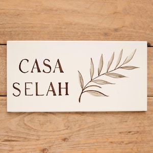 Op de afbeelding: Een rechthoekig, crèmekleurig bord met de woorden "CASA SELAH" in donkerbruine letters. Een delicate, handgeschilderde tak met bladeren in grijstinten en bruin siert de rechterkant. Het bord ligt op een houten oppervlak.