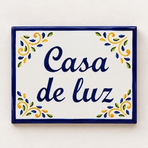Könnte beinhalten: Eine rechteckige Keramikfliese mit weißem Hintergrund und dunkelblauem Rand. Der Text Casa de luz ist mittig in einer dunkelblauen Schreibschrift aufgedruckt. Jede Ecke ist mit einem floralen Muster in Gelb, Grün und Blau verziert.