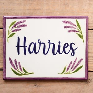 May include: A white ceramic sign with a purple border and lavender floral accents. The sign says "Harries" in blue cursive lettering.