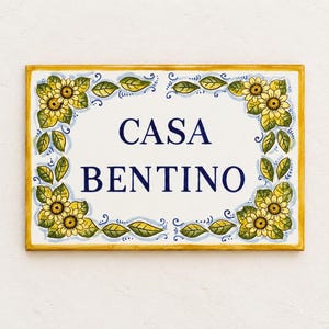 Könnte beinhalten: Ein rechteckiges Keramik-Hausschild mit weißem Hintergrund und gelbem Rand. Der Text CASA BENTINO ist in dunkelblauen Großbuchstaben in der Mitte aufgedruckt. Der Rand ist mit handgemalten gelben Sonnenblumen und grünen Blättern verziert.