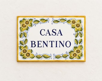 May include: A rectangular ceramic house sign with a white background and a yellow border. The text CASA BENTINO is printed in dark blue capital letters in the center. The border is decorated with hand-painted yellow sunflowers and green leaves.
