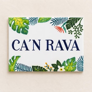 Puede incluir: Un letrero rectangular de cerámica blanca con el texto en azul oscuro CA'N RAVA centrado en la superficie. El borde está decorado con una ilustración pintada de varias hojas tropicales verdes, pequeñas bayas amarillas y una flor blanca.