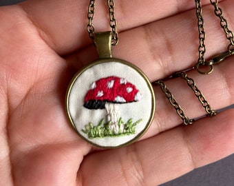 May include: A gold chain necklace with a round pendant featuring an embroidered red and white mushroom with black spots on a white background.