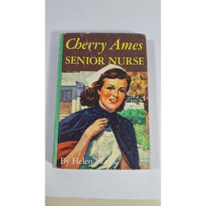 May include: Vintage book cover for "Cherry Ames, Senior Nurse." The cover features a smiling woman in a nurse's uniform and cap, with a dark blue cape. The title is in yellow, and the author's name is visible.