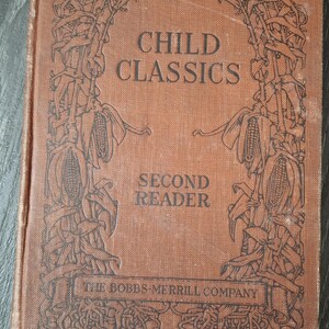 May include: A vintage brown book with the title "CHILD CLASSICS SECOND READER" and the publisher "THE BOBBS-MERRILL COMPANY" printed on the cover. The cover features an ornate design of corn stalks.