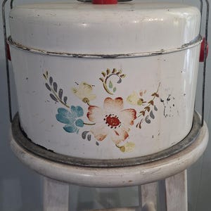 May include: A round white metal cake carrier with a red knob on the lid and a metal swing handle. The exterior features a painted floral design with blue, orange, and green leaves and petals. The metal shows signs of wear and paint chipping.