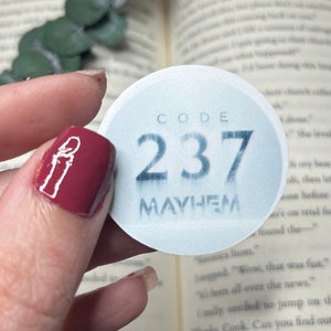 Op de afbeelding: Een ronde witte sticker met blauwe tekst die "CODE 237 MAYHEM" zegt. De sticker wordt vastgehouden door een hand met rode nagellak.