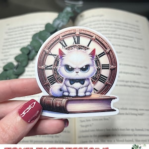 Op de afbeelding: Een witte kat met een strik zit op een boek met een klok erachter. De kat heeft een boze uitdrukking. Het boek is dicht en heeft een versleten leren kaft. De wijzerplaat is goudkleurig met Romeinse cijfers. De sticker is voor een stickercollectie.