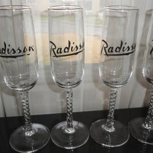 May include: Four clear glass champagne flutes with a twisted stem. Each flute has the word "Radisson" printed in black on the side.