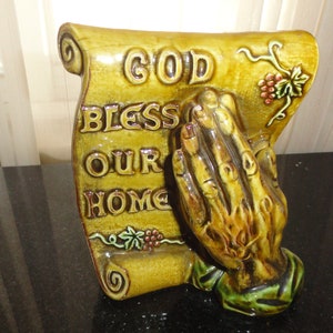May include: A ceramic figurine of a praying hand holding a scroll with the words "God Bless Our Home" in brown lettering on a yellow background. The scroll is decorated with green vines and red grapes.