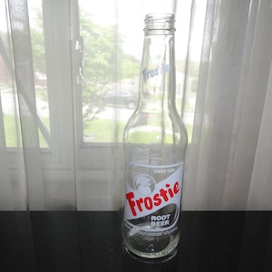 May include: A clear glass bottle with a red and white label that says "Frostie Root Beer" and "Since 1923".