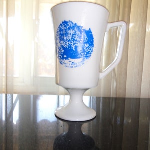 May include: White ceramic coffee mug with a blue illustration of a forest scene and the text "Mary's Rock Tunnel Skyline Drive VA". The mug has a pedestal base.