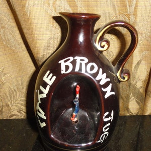May include: A brown ceramic jug with a white inscription that reads "Little Brown Jug". The jug has a handle and a cutout in the front.