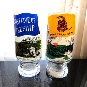 May include: Two clear glass tumblers with a wide base. One features the text "DON'T GIVE UP THE SHIP" on a blue background, with a ship illustration. The other has a yellow background with a coiled snake and the text "DON'T TREAD ON ME".