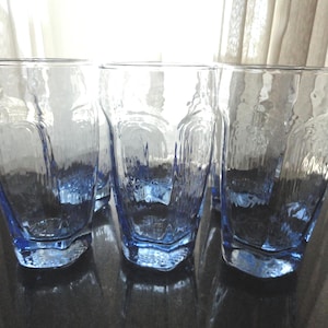 May include: Set of four clear drinking glasses with a textured surface and a blue tint at the base. The glasses have a unique, irregular shape, and are suitable for water, juice, or other beverages. The glasses are displayed on a dark surface.