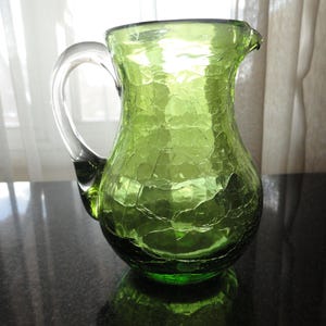 May include: A green glass pitcher with a clear handle and a spout. The pitcher has a crackle finish, giving it a textured appearance. The pitcher is sitting on a dark surface.