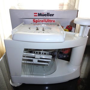 May include: White Mueller SpiralUltra vegetable spiralizer with a clear container and a white tray. The box behind it has the Mueller logo and product name. The spiralizer is designed for easy vegetable preparation.
