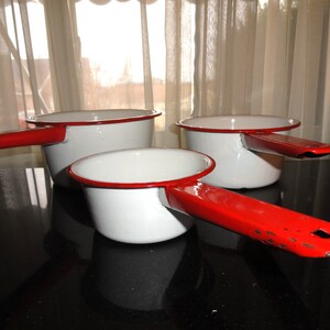 May include: Set of three white enamel saucepans with red rims and handles. The saucepans are nested together on a black surface.