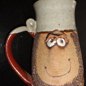 May include: A handmade ceramic mug with a whimsical face design. The mug features a tan body with a smiling face, white eyes, and a red handle. The top of the mug is a light beige color.